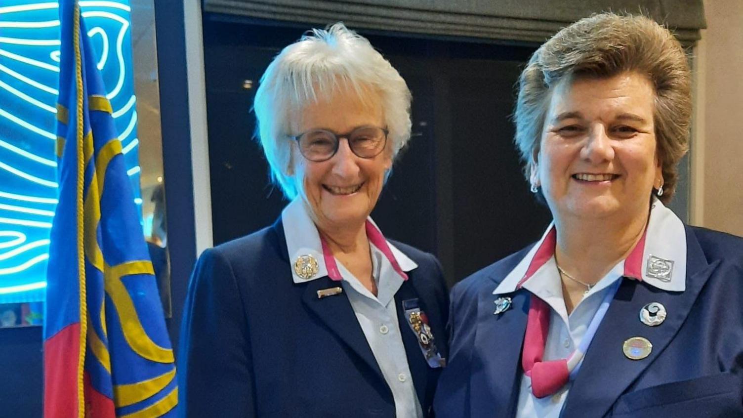 Two older women with short hair wearing light blue shirts and navy blazers, the uniform of Girlguiding UK volunteers.