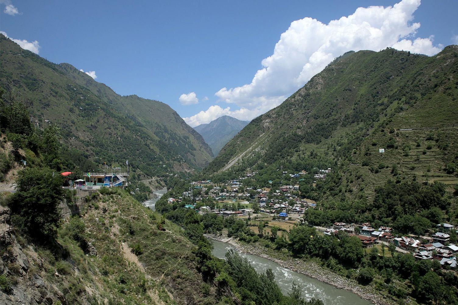 A valley scene with a river winding through lush green mountains. A small village with scattered houses lines the riverbanks.