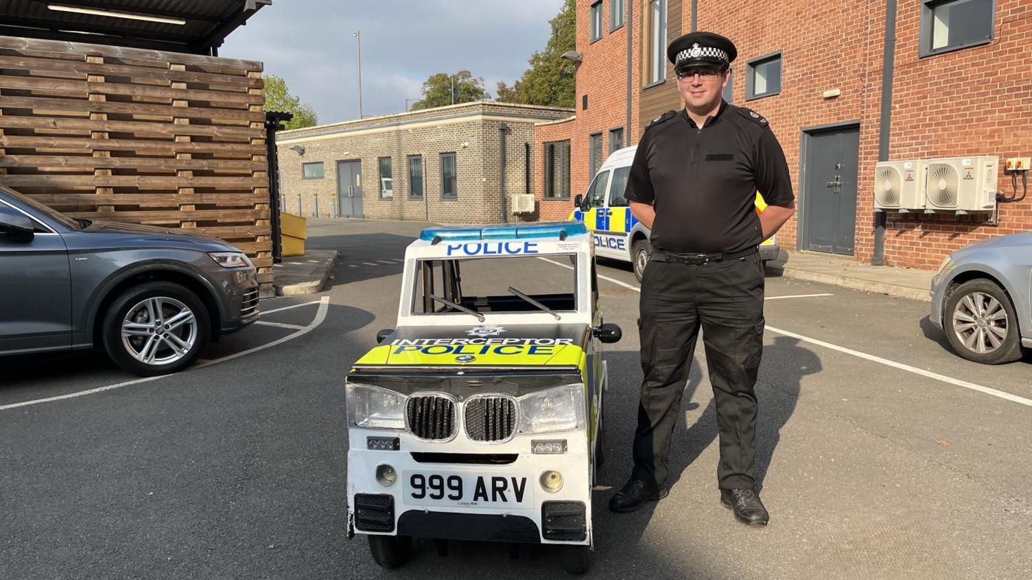 PC Daniel Brock wearing a black police officers uniform and standing next to a soap box cart that resembles a police car