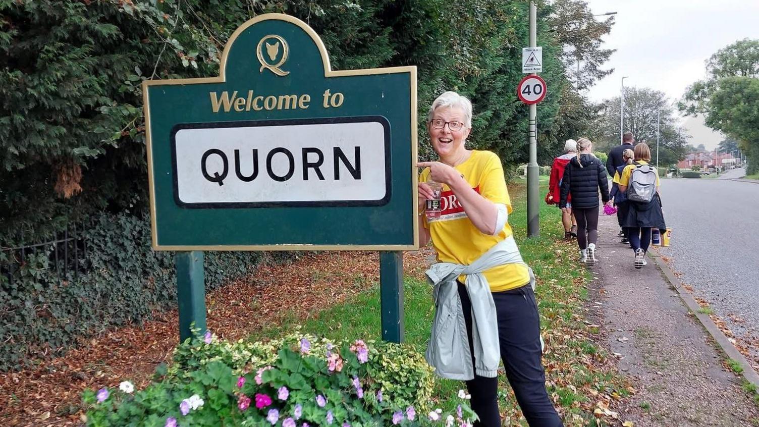 Laura stood in a yellow t shirt pointing at a sign saying welcome to Quorn and smiling.
