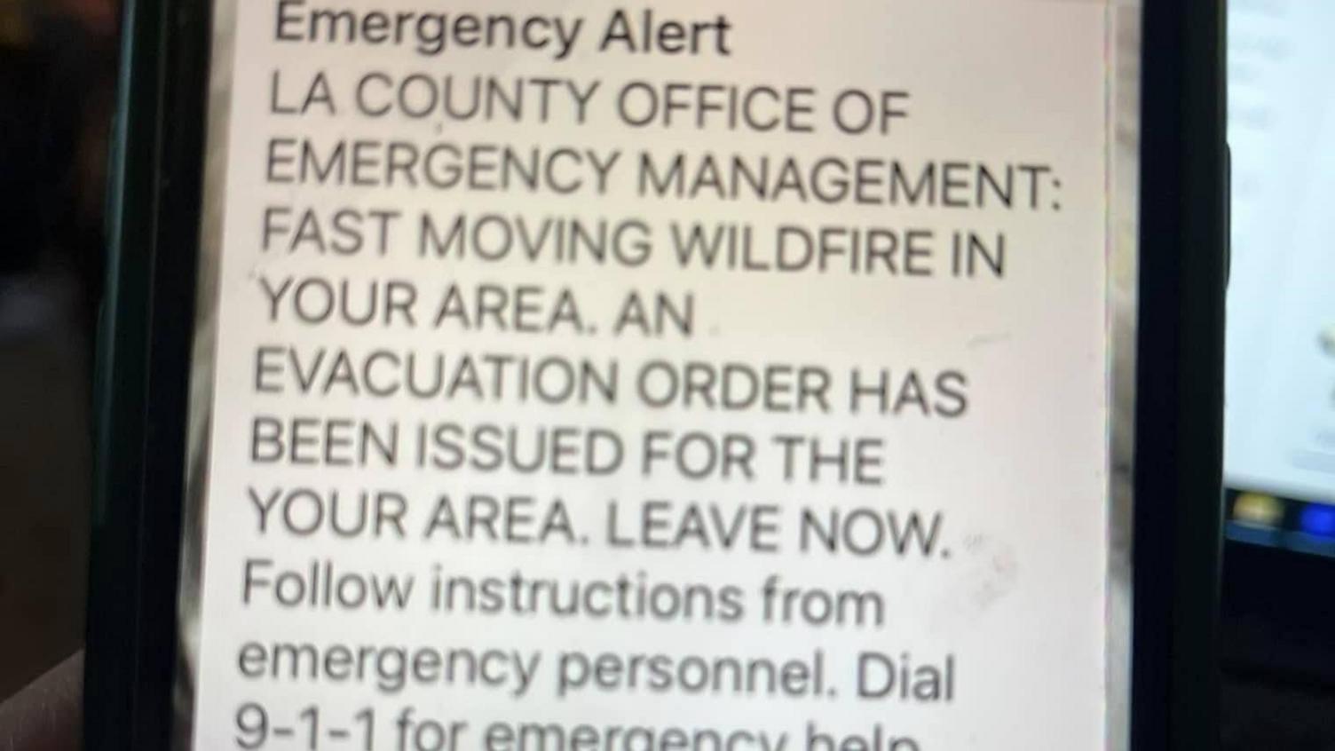 A mobile phone with a text message reading:  Emergency Alert.  LA COUNTY OFFICE OF EMERGENCY MANAGEMENT: FAST MOVING WILDFIRE IN YOUR AREA.  AN EVACUATION ORDER HAS BEEN ISSUED FOR YOUR AREA.  LEAVE NOW.  Follow instructions from emergency personnel.  Dial 9-1-1 for emergency help. 