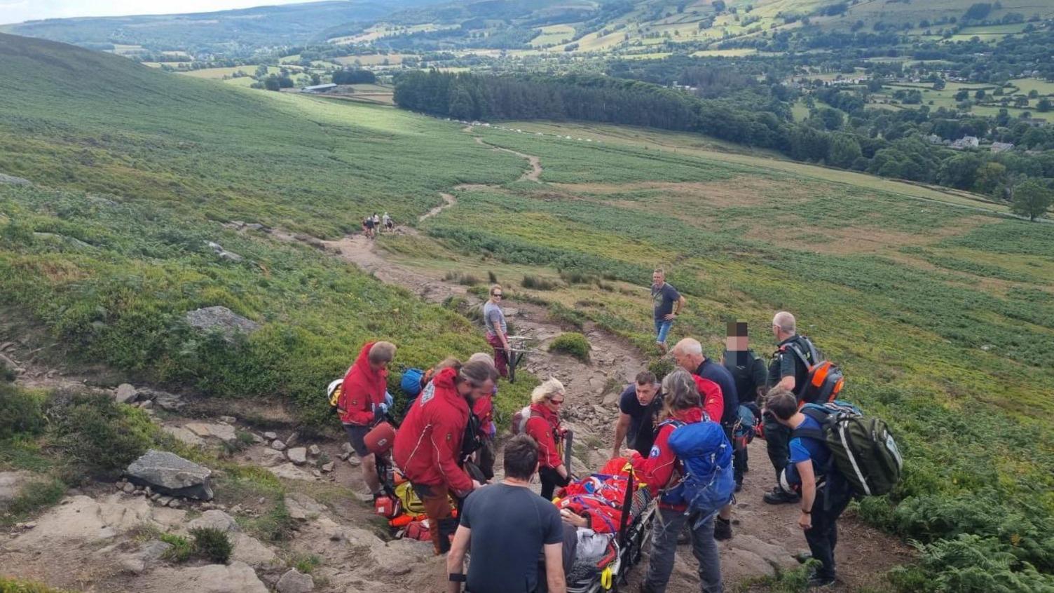 A team of volunteers in outdoor clothing carry a person a stretcher down a path towards trees at the bottom of a valley