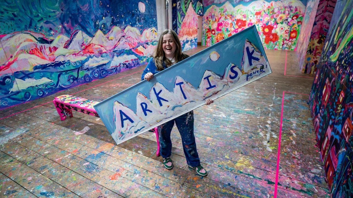 Emily Powell pictured holding a large sign reading "Arktisk" with mountains painted on it. 