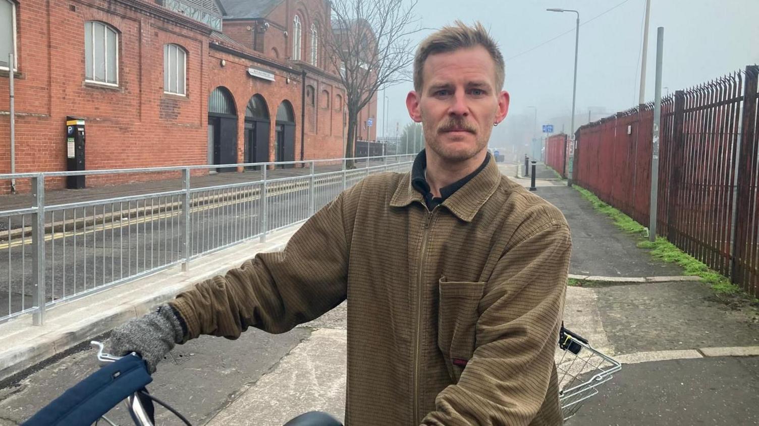 A man wearing a brown over shirt is standing in a bike lane holding his bicycle handlebars to one side. There is a basket on the back of the bike. He has fair hair and a brown moustache. 