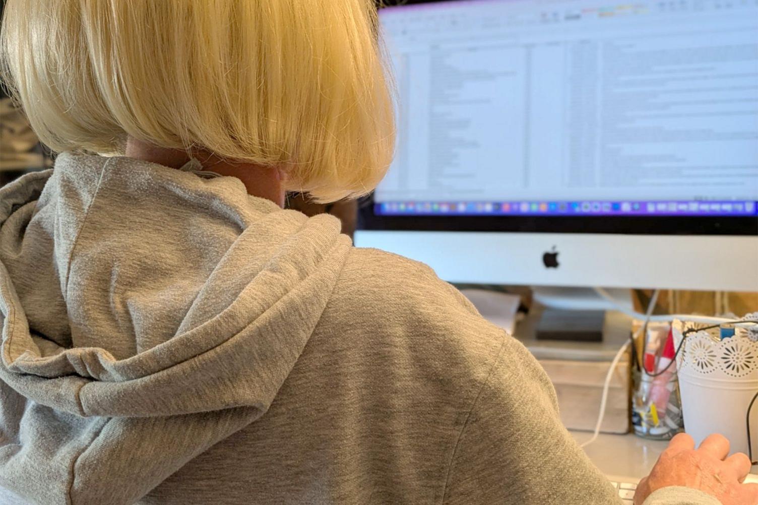 Gemma sits at a desk facing a large computer monitor displaying a spreadsheet or document with multiple columns and rows. She is facing away from the camera, and her face cannot be seen, with blonde hair and a grey hoodie. On the desk, there is a white mug with decorative patterns and a clear container filled with pens and pencils.
