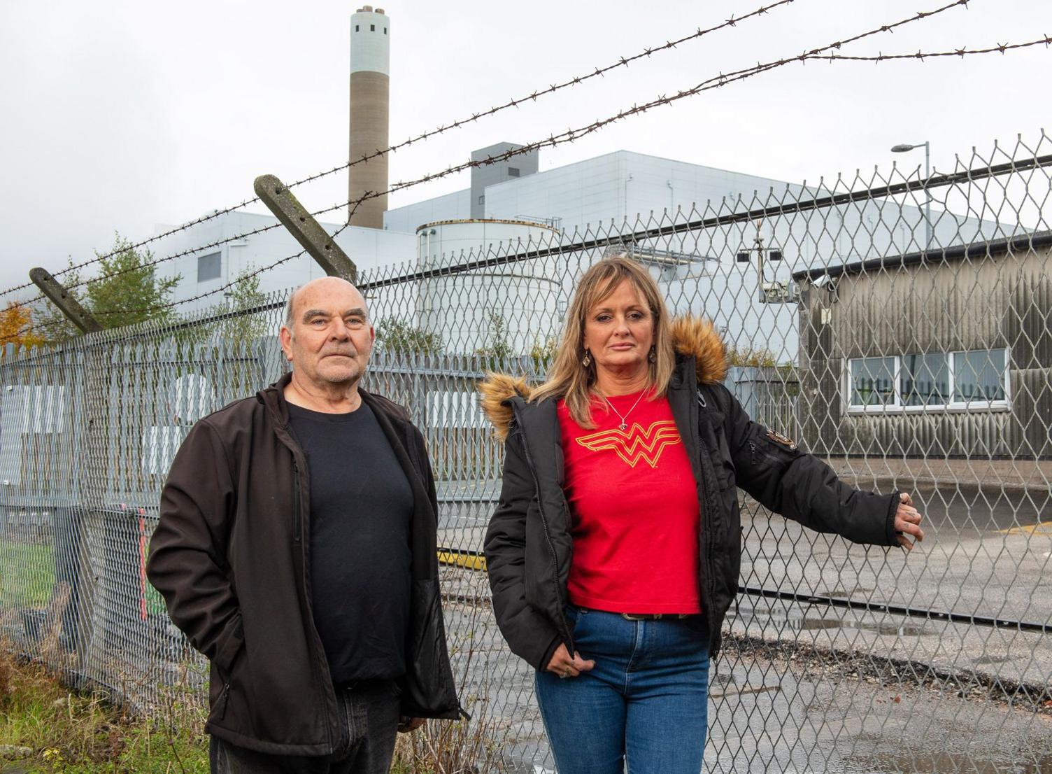 Runcorn residents George Parker, garage owner and Mandy Royle stand solemnly infront of a fence. In the background is the Runcorn waste incinerator, which backs onto their properties.   