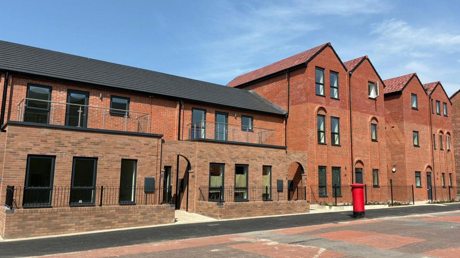 A row of modern terraced properties with black metal railings. A tarmac pavement runs along the front and there is a red post box.