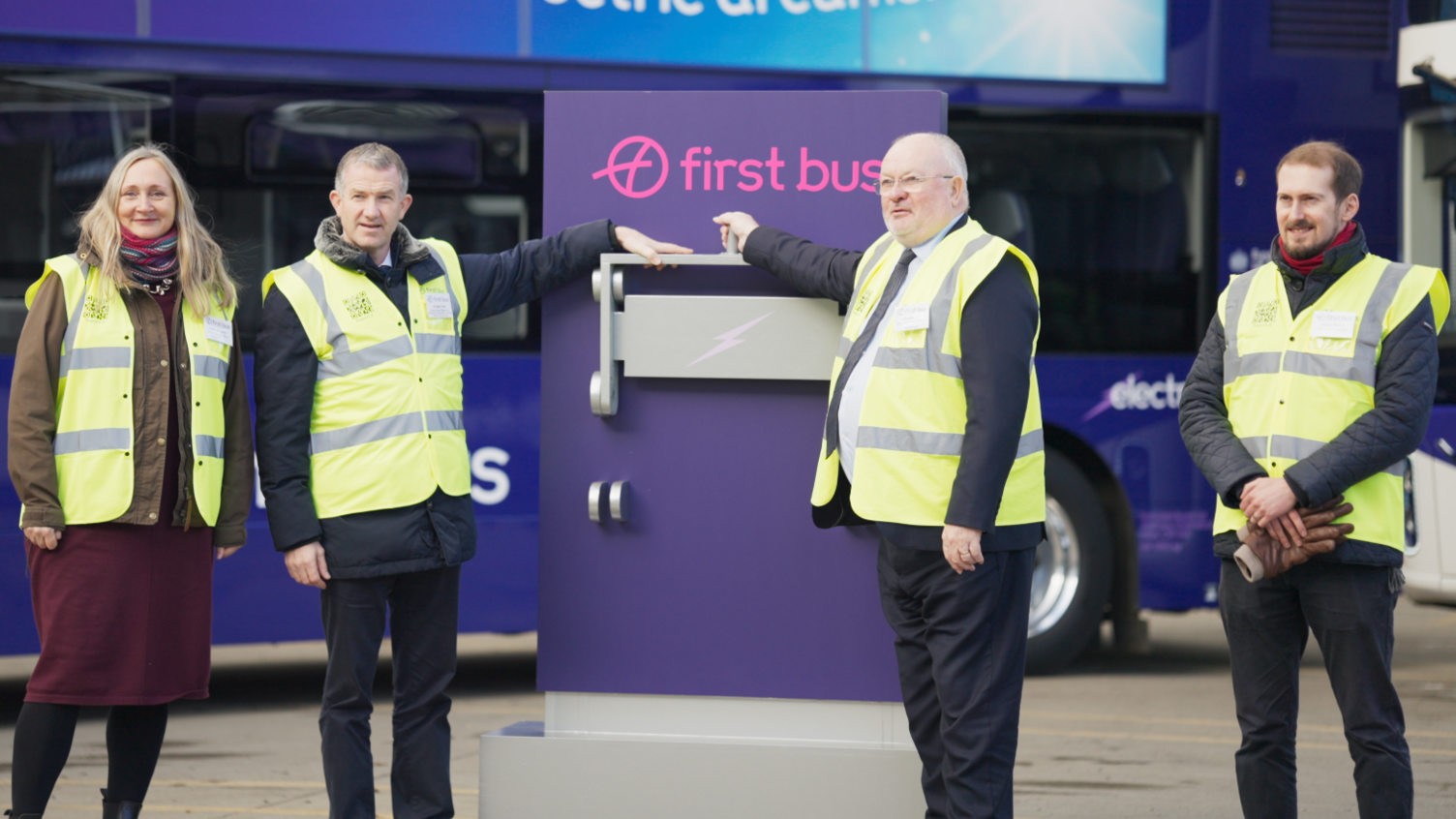 A woman and three men are standing in yellow fluorescent tabards. In the background are buses. They are all looking at, or slightly to the left, of the frame.