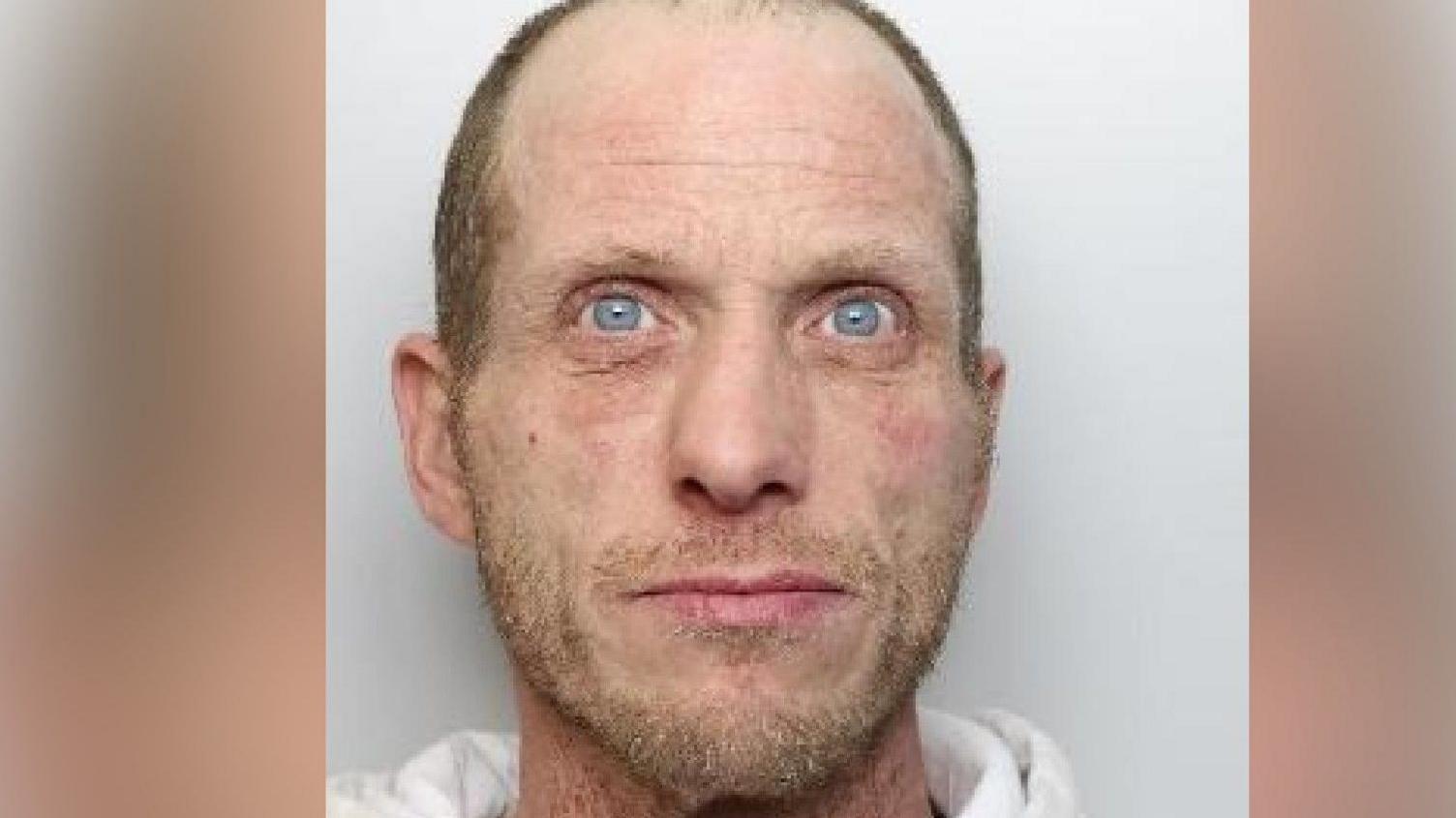 A mugshot of a man who is wearing a white jumper. He has blonde facial hair and short, shaven hair which reveals a receding hairline.