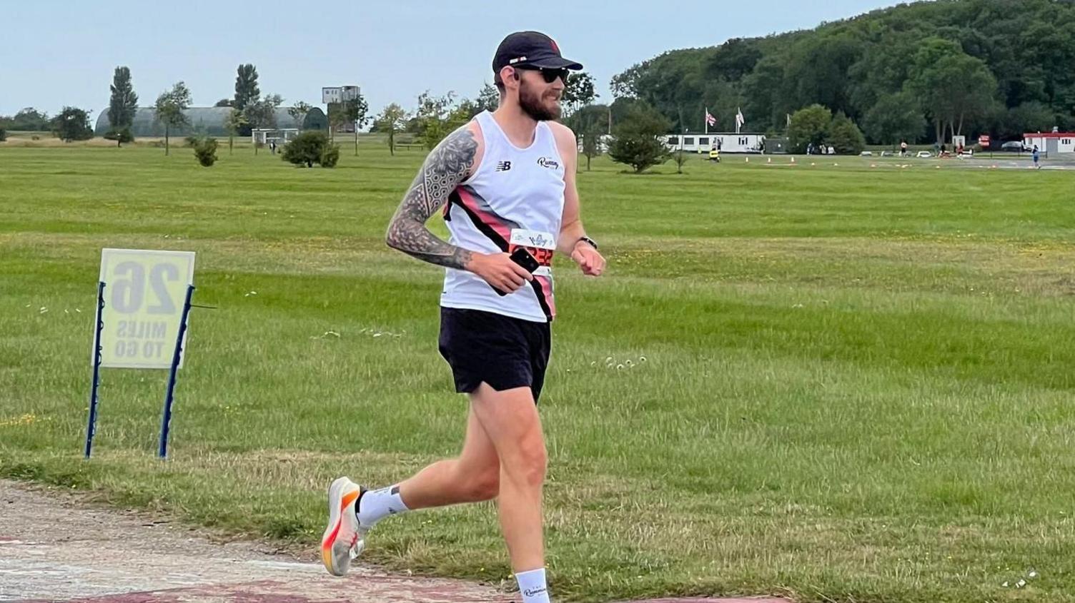 Alex with short dark hair and beard, dressed in running kit, running past a large area of grass with trees and a mountain in the background. He has just passed a 26 mile marker.