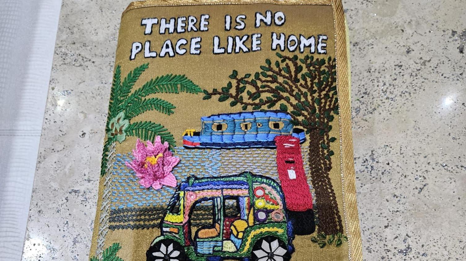 An embroidered book cover with the words there is no place like home at the top. There is a palm tree, a red post box, a tuktuk taxi, a waterlilly and a canal boat