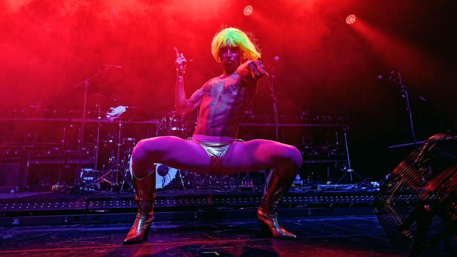 Shanika Sunrise wearing a yellow wig, metallic boots and pants. They are standing on a stage