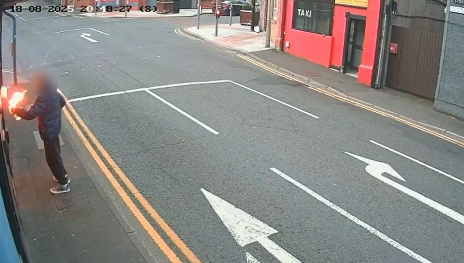 A CCTV grab of Irish Street, a wide road with a red building on the corner to the right with a sign saying "Taxi" in the window. To the left of the frame a figure in a blue puffy jacket and dark tracksuit bottoms can be seen holding a flaming object.