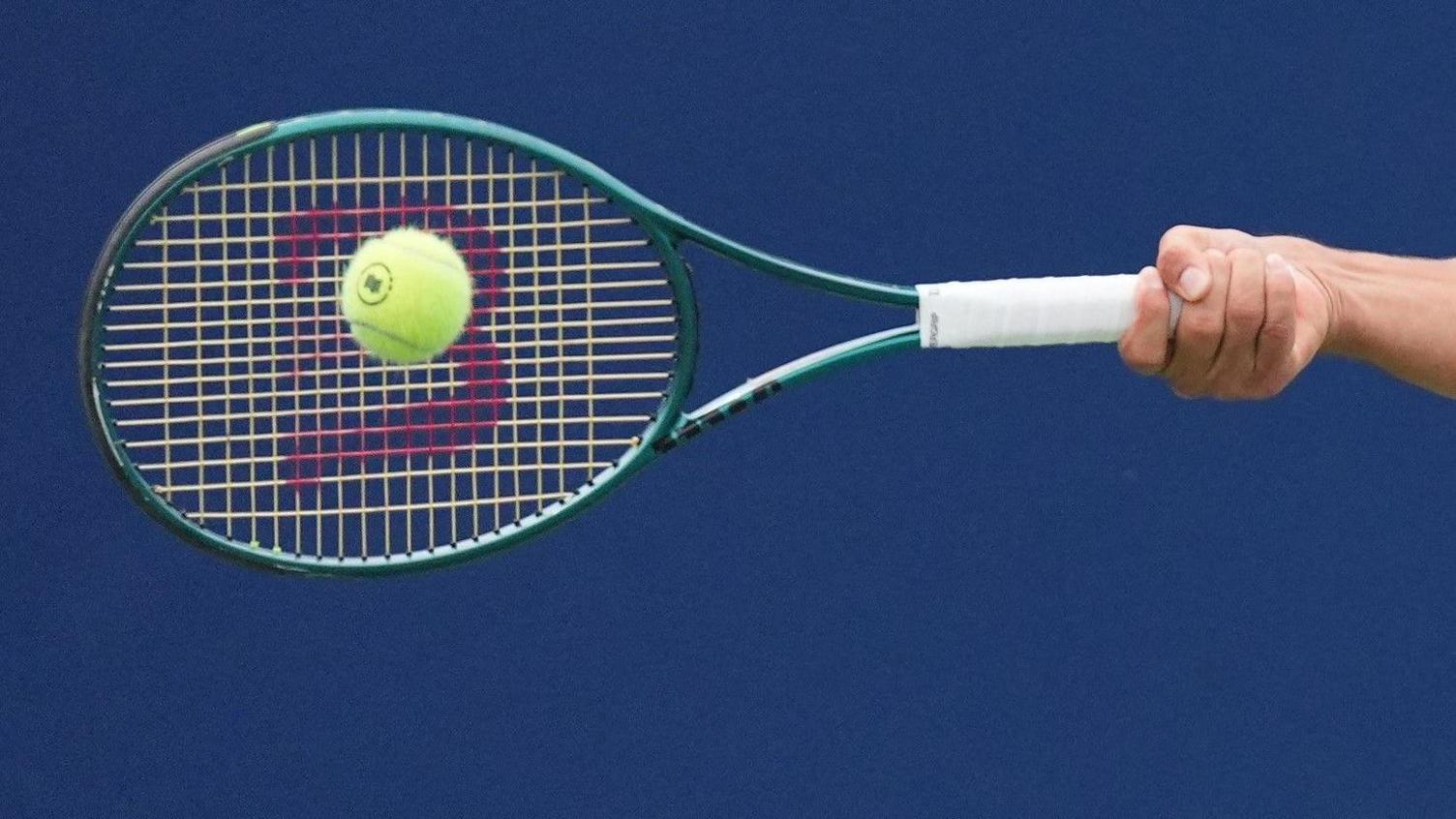 A tennis racket, against a dark blue back drop, is hitting a tennis ball.
