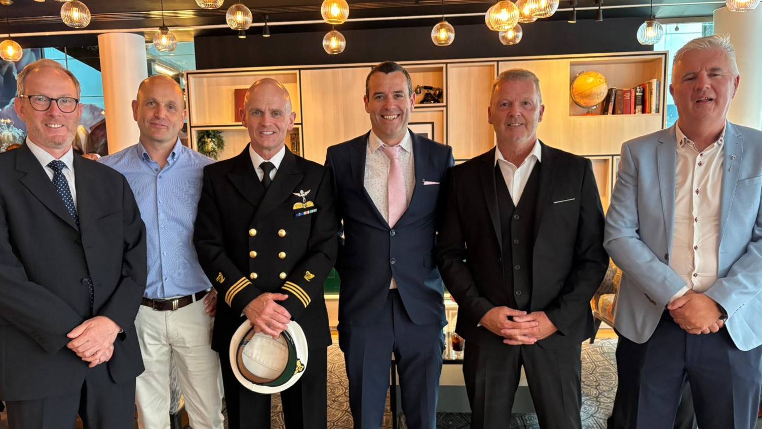 The image shows six men standing side by side facing the camera. They are all dressed in suits, bar one who is in a uniform. 
