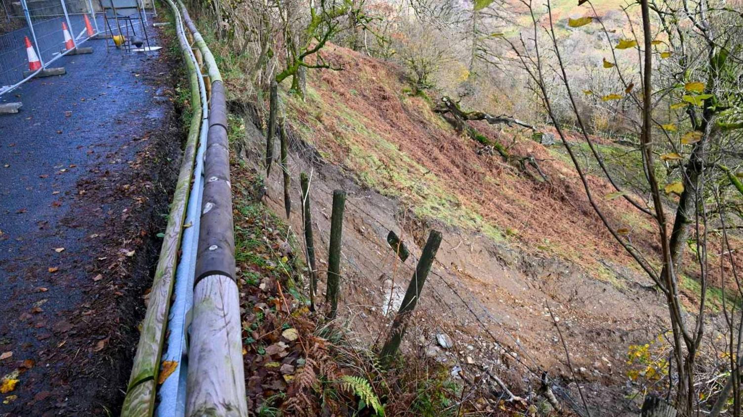 A picture taken from the road looking down at the landslip. Fence posts are dangling in the air above a track of brown, wet soil.