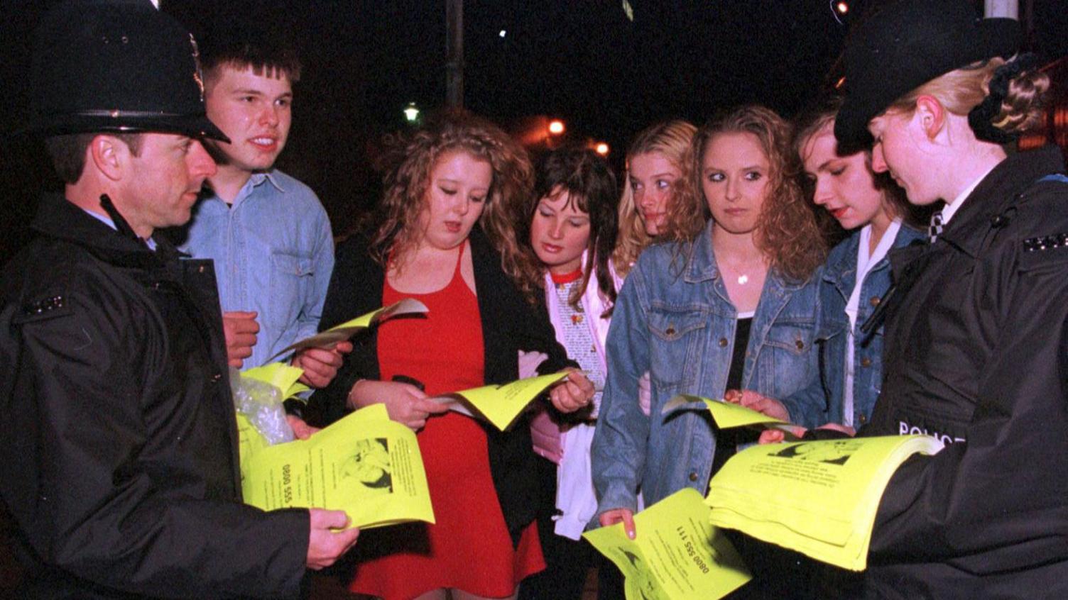 Two police officers handing out yellow posters to a group of young people outside a nightclub. The posters have a picture of Leah Betts in hospital on them and warn of the dangers of taking drugs.