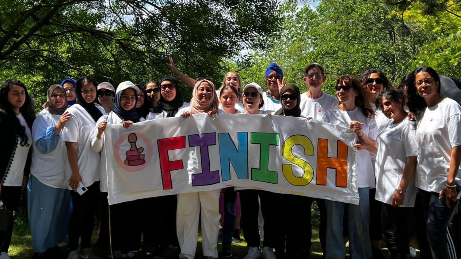 A group of 20 women lined up across the photo. They are holding a sign that reads "finish", and appear to have completed a fundraising event. They are all wearing white T-shirts. They are stood with lots of trees in the background behind them.