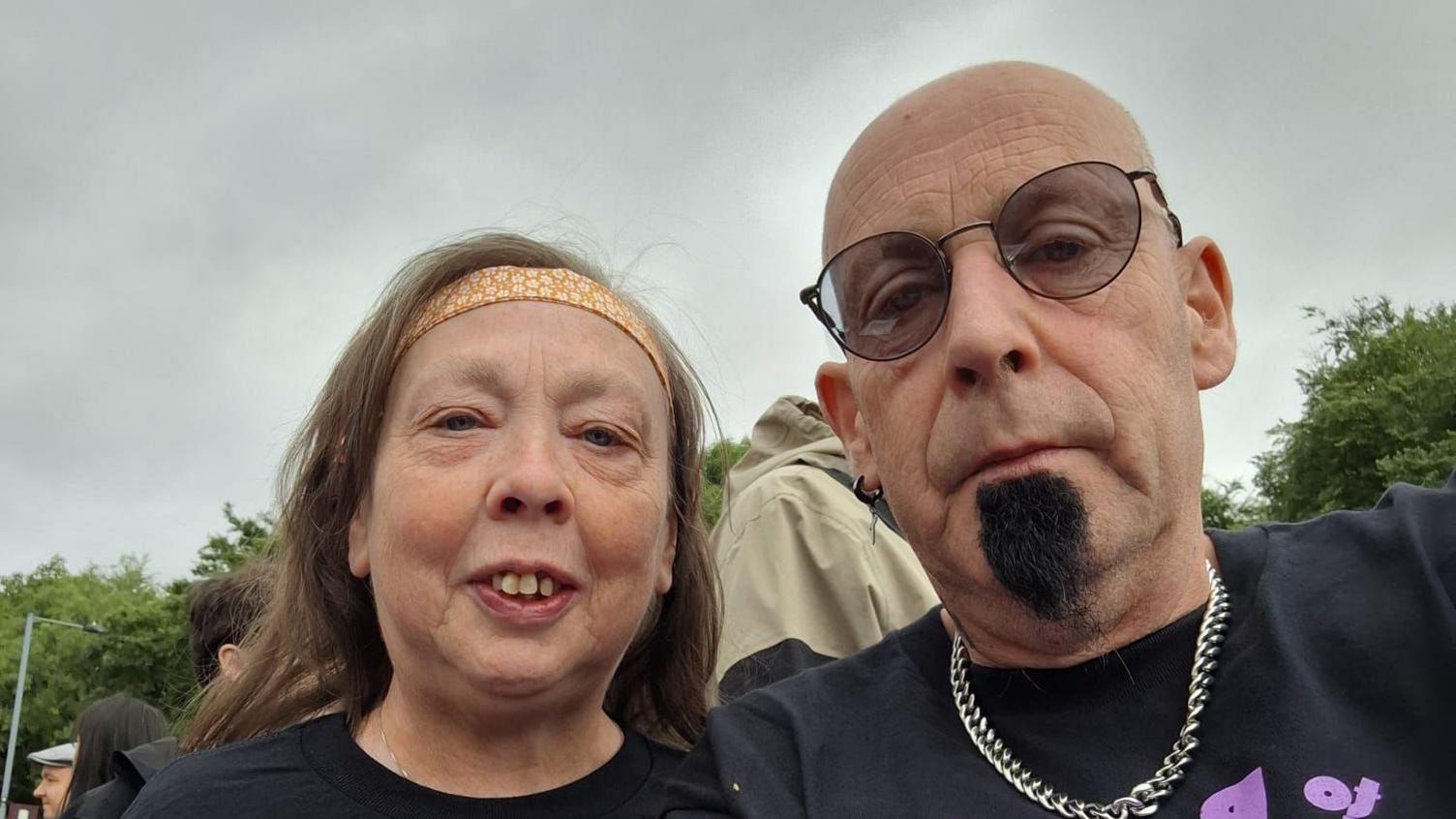 A man and a woman who are wearing Black Sabbath concert t-shirts looking at the camera. The man is bald and has a black goatee. The woman has brown hair and is wearing an orange headband 