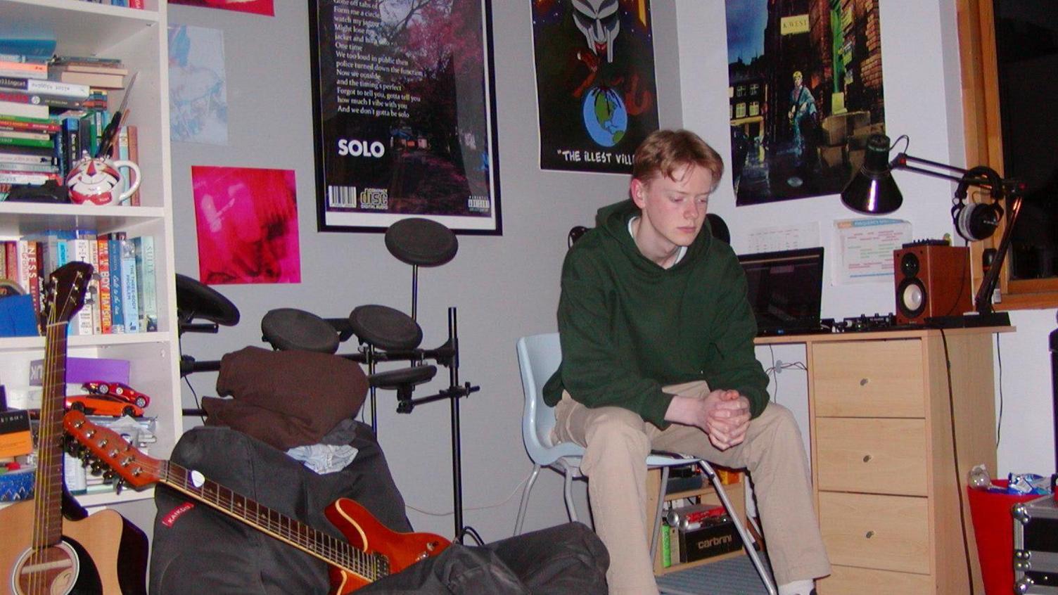A man with parted red hair sits on a chair in the corner of a room with his hands clasped. He is wearing tan trousers and a green hoodie. A laptop and speaker can be seen on the wooden desk behind him. A guitar sits on a grey bean bag, another rests beside it in front of a stacked bookshelf. There is also a drum set behind the bean bag and various posters on the walls of the room.