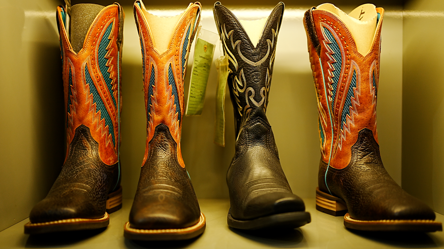 A row of three cowboy boots in orange, back and blue. They are in a box, facing forward. They are patterned on both the leather and the material tops