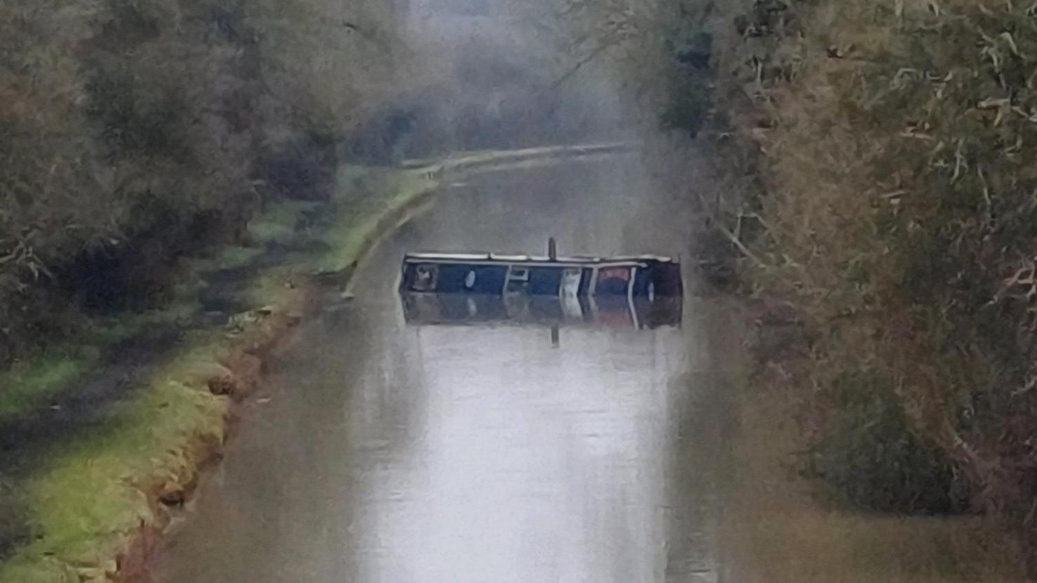 A canal with a partially-submerged barge positioned across it. A tow path is on the left of the canal with trees either side of the waterway. The canal curves to the right and out of sight in the distance.