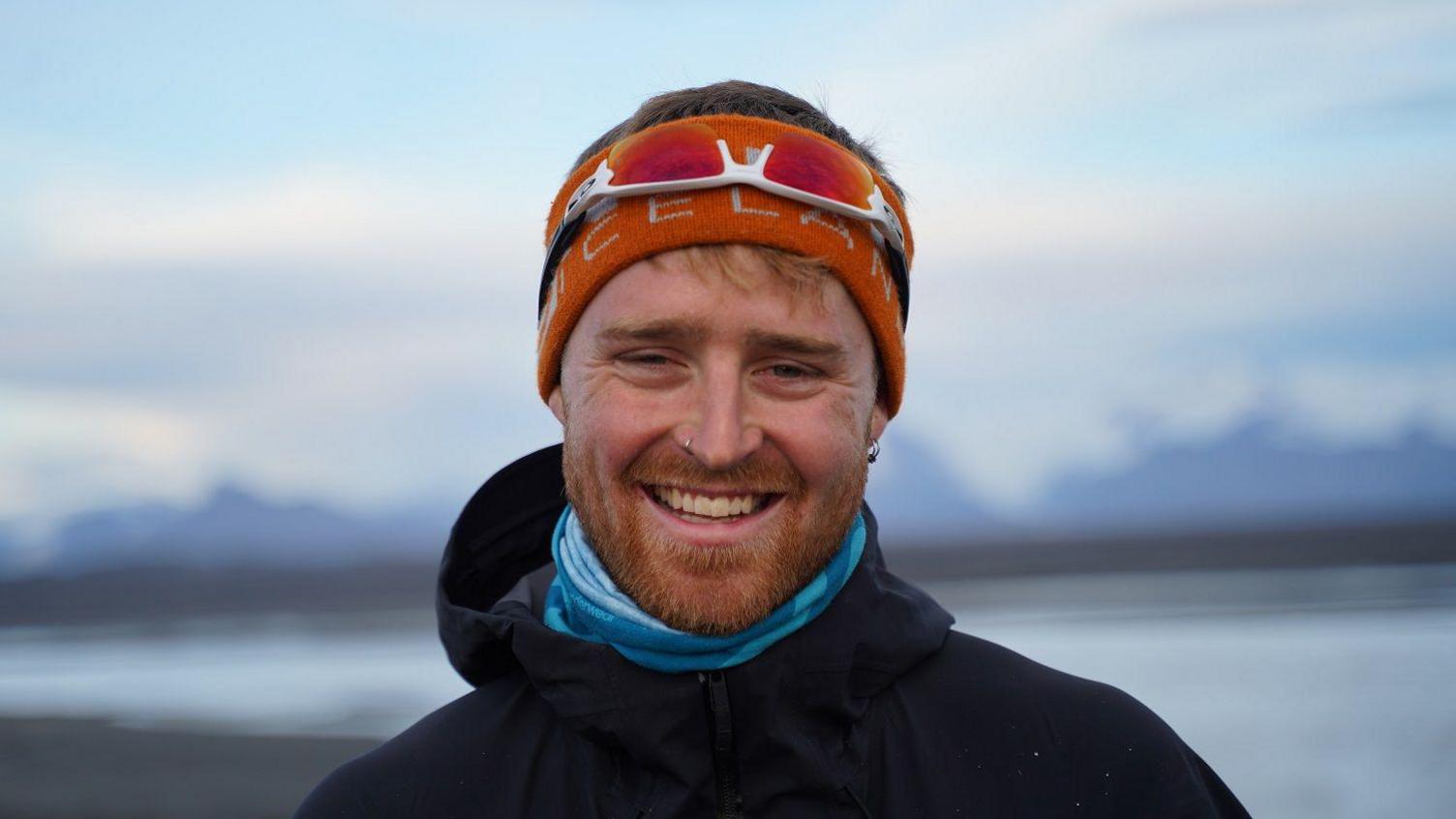 A man wearing an orange headband and a pair of sunglasses on his head, and a black waterproof coat with a blue gaiter, smiling at the camera with a snowy landscape in soft focus behind him. He's got stubble on his face and is wearing an earring and a nose ring.