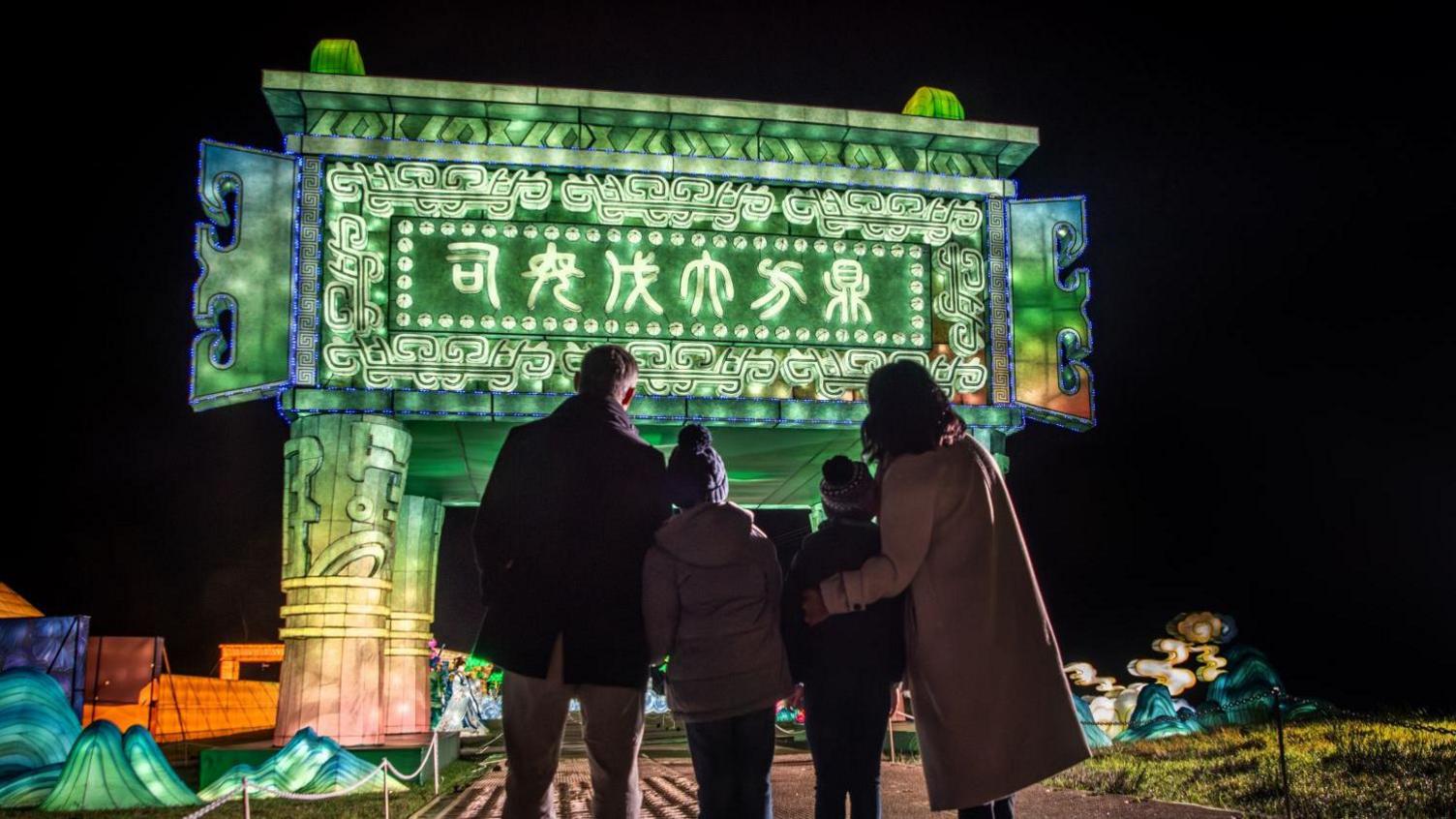 A family of two parents and two children stand and look at the giant welcome arch at the Longleat Festival of Light. It is glowing green and has Chinese writing on it. The picture is taken at night and other illuminations can be seen in the background