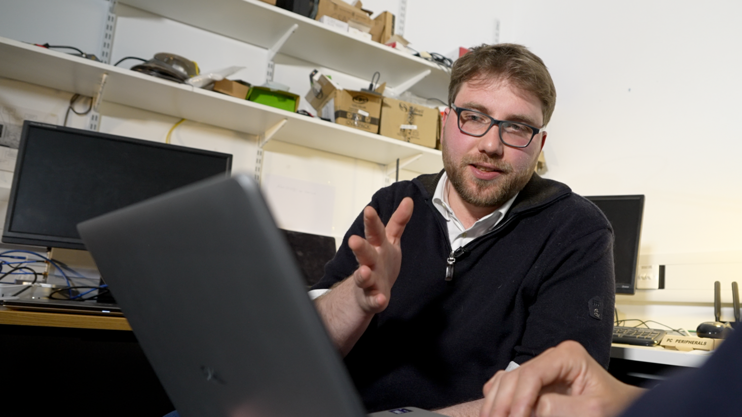 Photo of Luca Arnaboldi looking at a laptop, holding his hand up. He has brown hair, a beard and is wearing glasses. Behind him are computer monitors and shelves with boxes. The room is painted white.