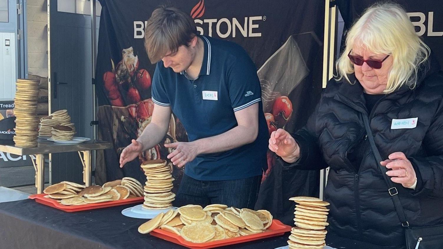 A man on the left is wearing a blue top, and is looking downwards at a stack of pancakes. The woman on the right is wearing a black jacket, and sunglasses, is stacking pancakes as well. 