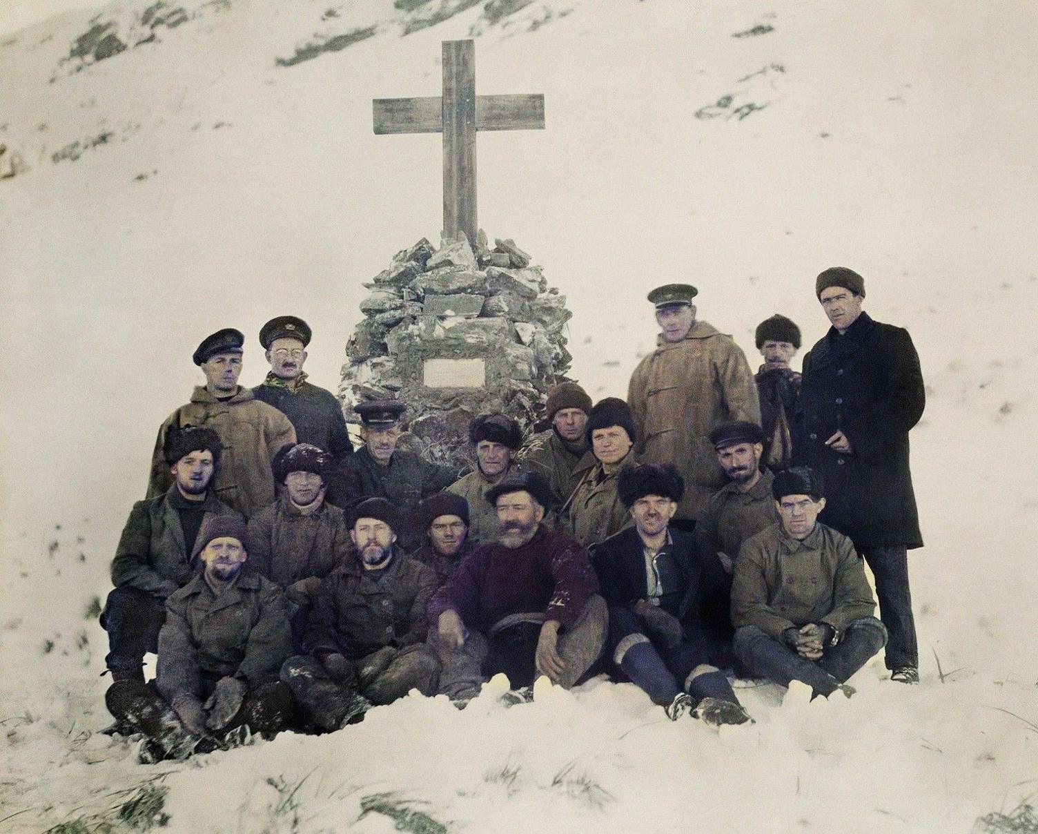 A group of 16 men from the crew of the Quest exploration ship, standing and sitting in front of a memorial cross to Sir Ernest Shackleton