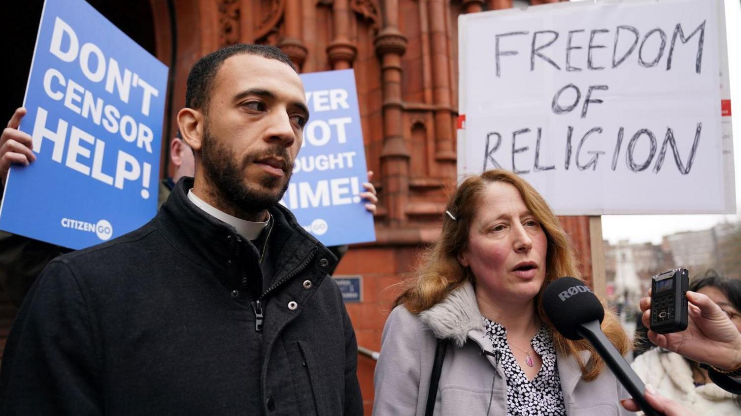 Father Sean Gough and Isabel Vaughan-Spruce outside Birmingham Magistrates' Court. Father Gough is wearing a black jacket and Ms Vaughan-Spruce is wearing a beige coat with a fur collar and black blouse with white spots. Behind Father Gough someone holds a placard saying 'Don't Censor Help!'. Another placard behind Ms Vaughan-Spruce says 'Freedom of Religion'. 