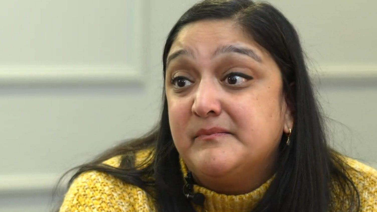 A woman with long black hair wearing a yellow knitted jumper looks at someone off-camera