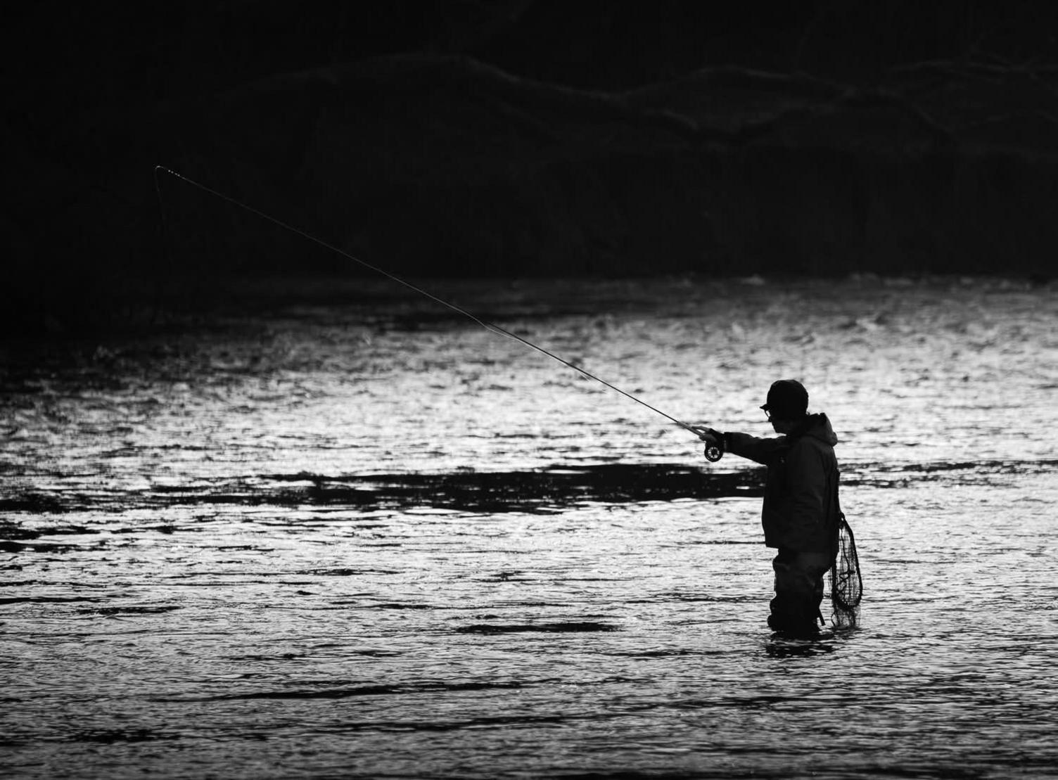 A loan fisherman with fishing rod in hand stands about knee deep in the River Tweed with a net behind him hoping to catch fish on a grey cold morning.