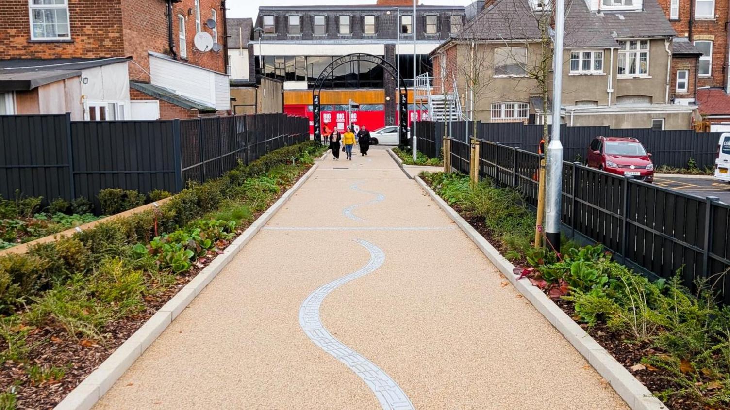 A large footpath heading towards an iron gateway with buildings in a town beyond it