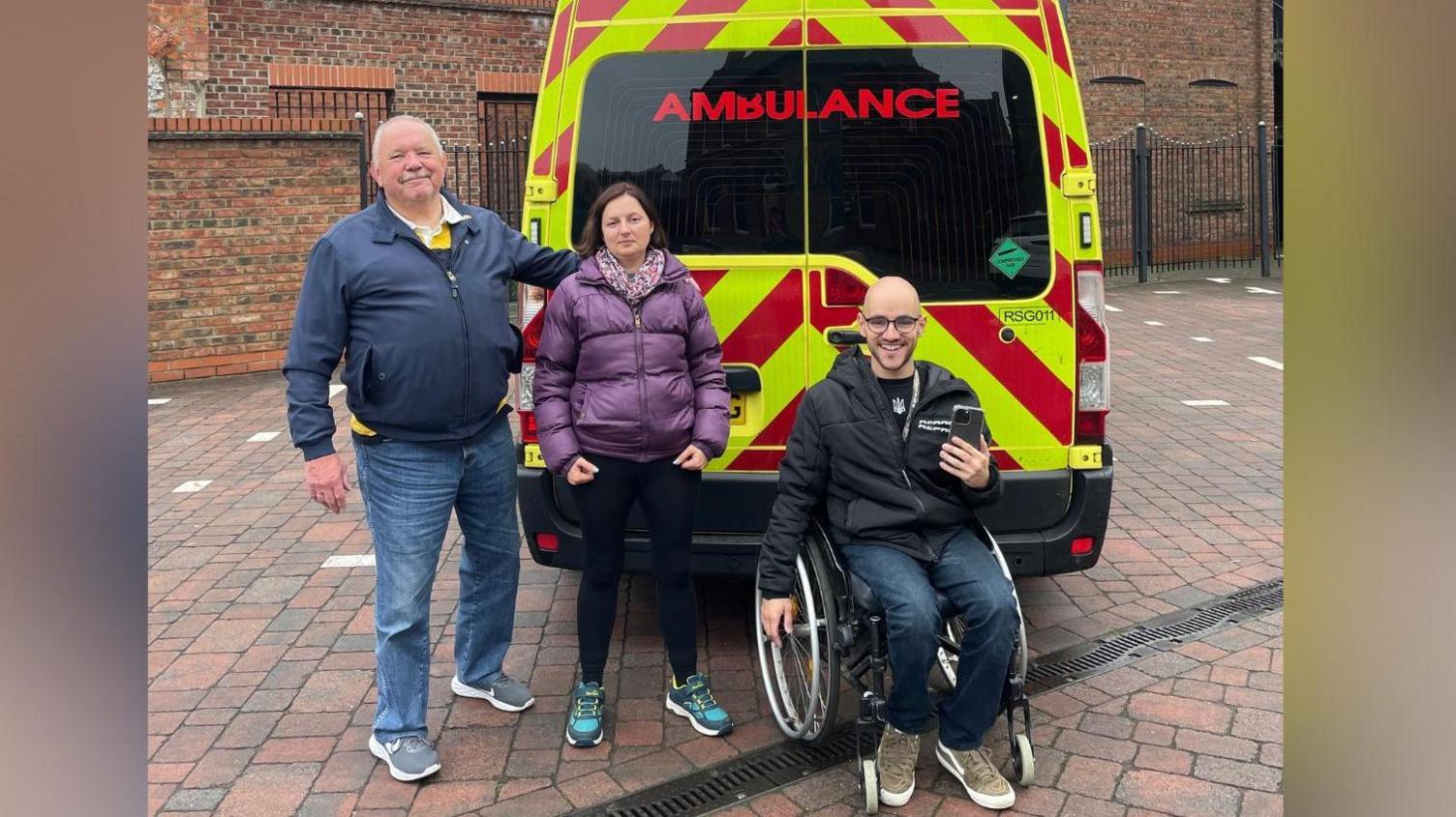 A man and woman stand in front of an ambulance in a courtyard, alongside a man in a wheelchair. The man standing to the left is wearing a blue coat and blue jeans and has grey balding hair. The woman is wearing a short purple coat and scarf and has short brown hair, while a man in a wheelchair has blue jeans and is wearing a short black coat. He has a bald head and is wearing black-rimmed glasses and is holding an iPhone in his left hand.