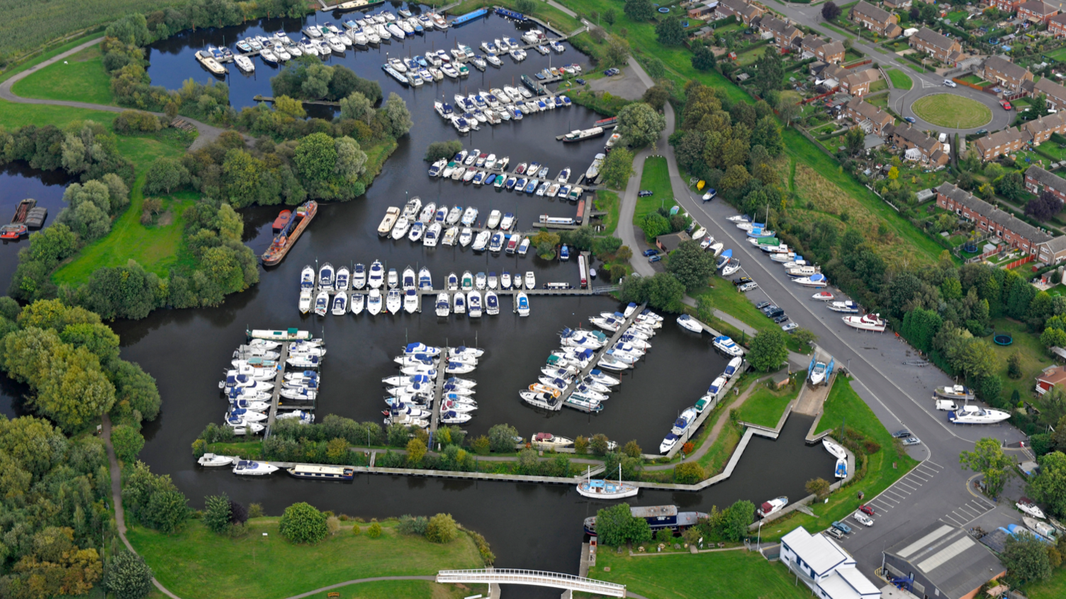 An aerial shot of a marina with many boats moored and houses on the left hand side of the image.