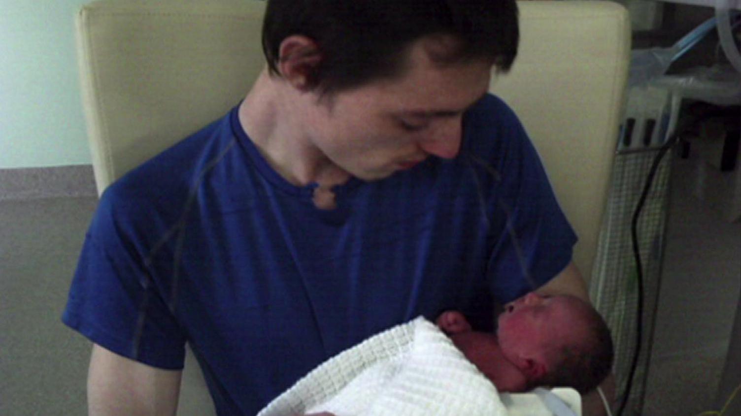 A new father holds a newborn baby in a hospital setting. He is wearing a blue T-shirt, with the baby swaddled in a white blanket.
