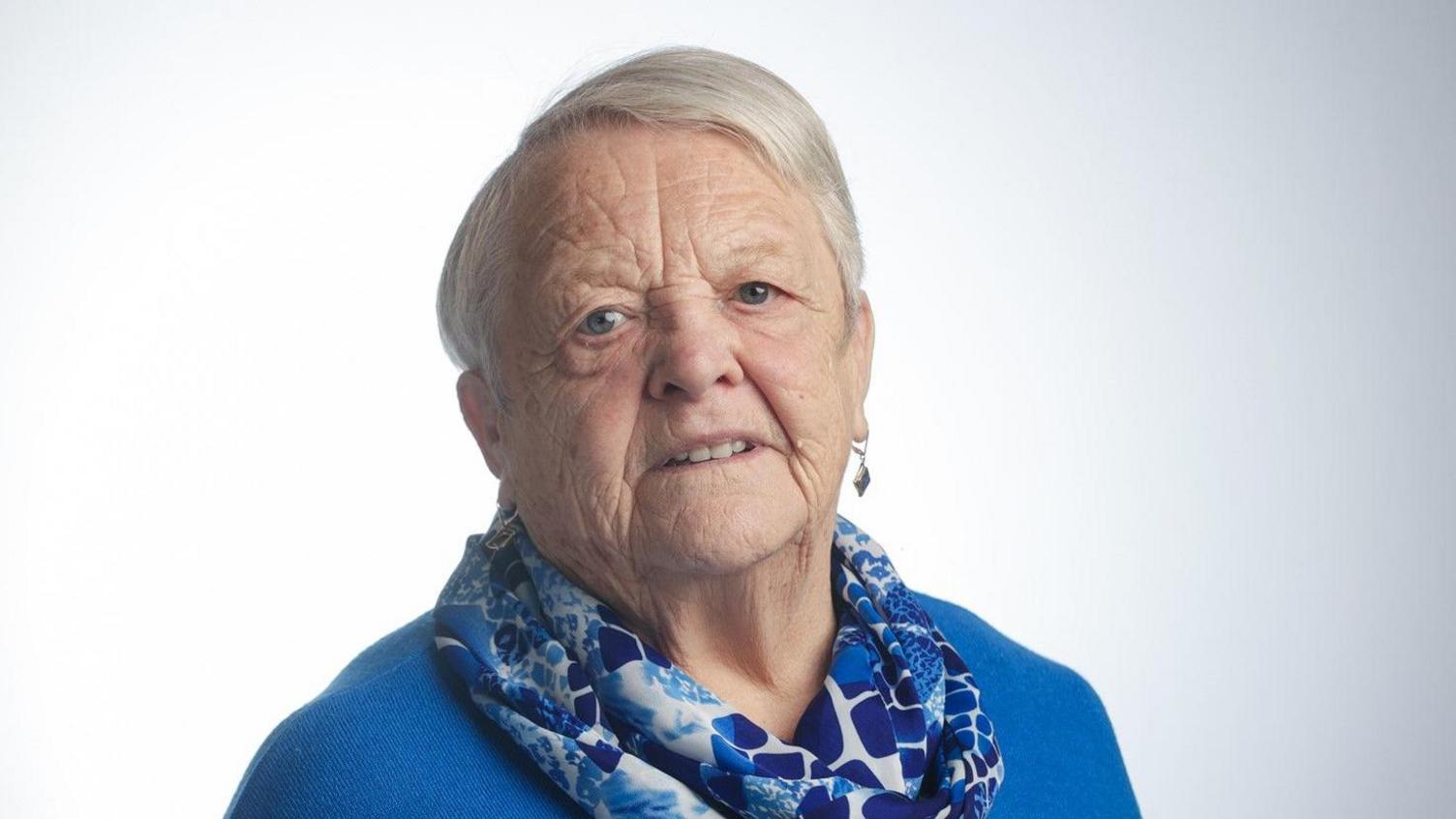 a woman against a blank background looking directly at a screen. She is wearing a blue top, a patterned blue and white scarf and drop earrings. Her white hair is styled short and swept over to one side