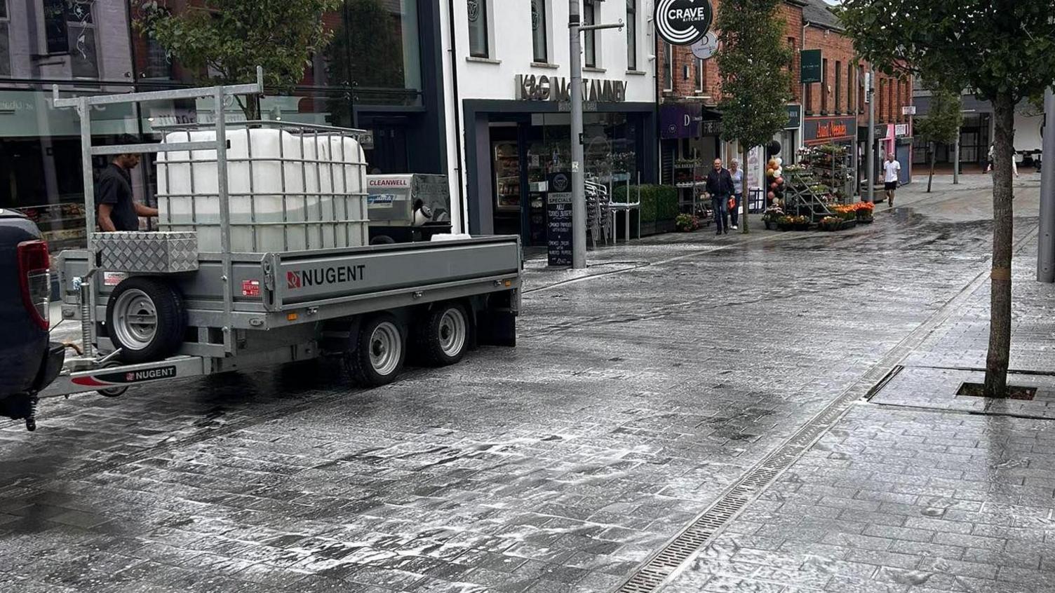 A black paved street with businesses down the side of it. There are suds on the ground as people clean up the slurry which was spread.