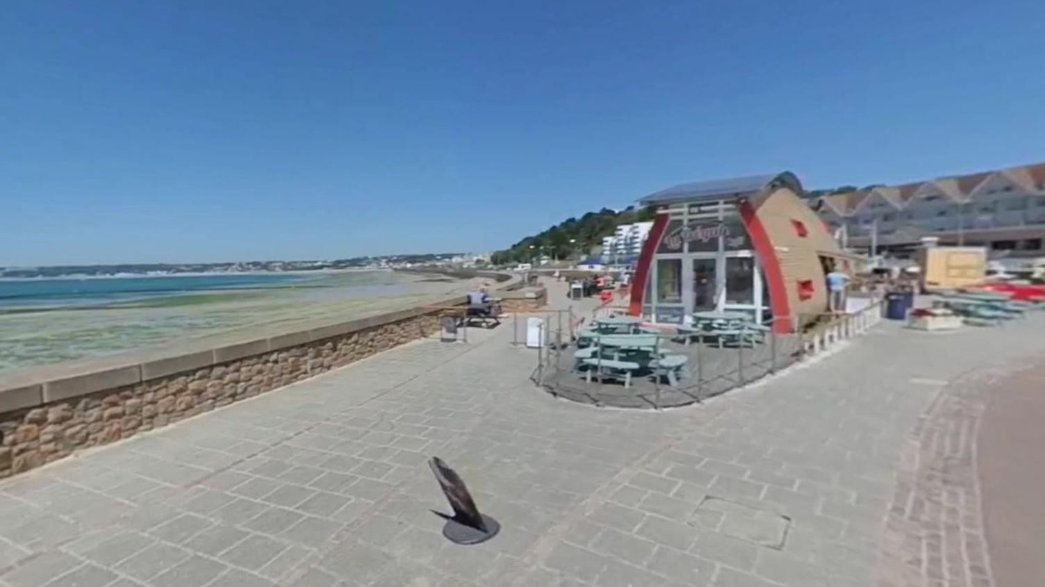 A picture of a café on a path next to the beach. A round building with glass windows and there is people sat on the tables outside of it.