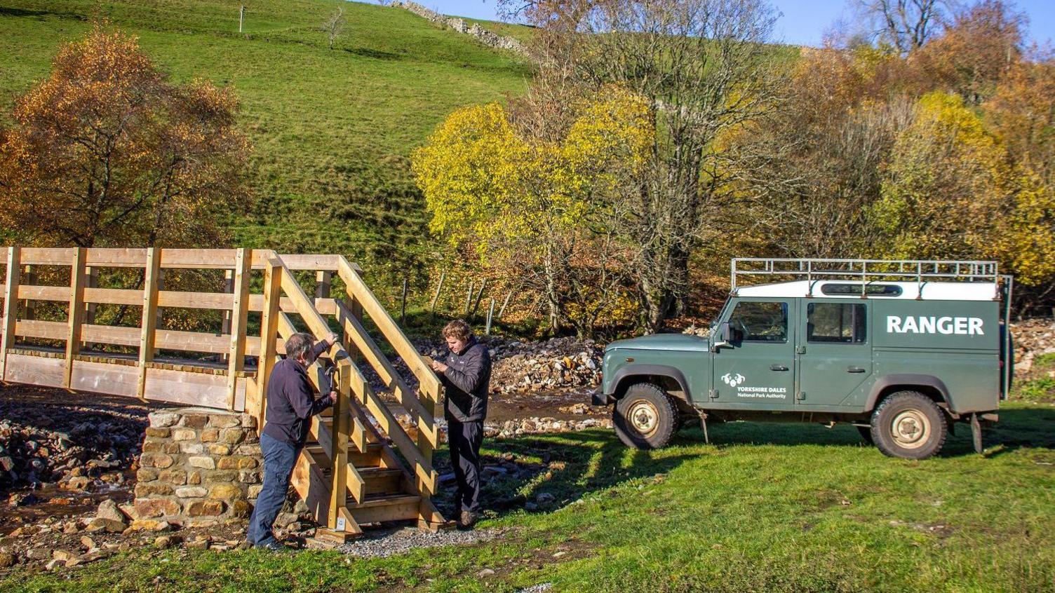 Two men fix a wooden bridge over a stream behind are green trees and hills. A dark green rangers 4x4 is parked behind them with the word 'RANGER' written in white on the side