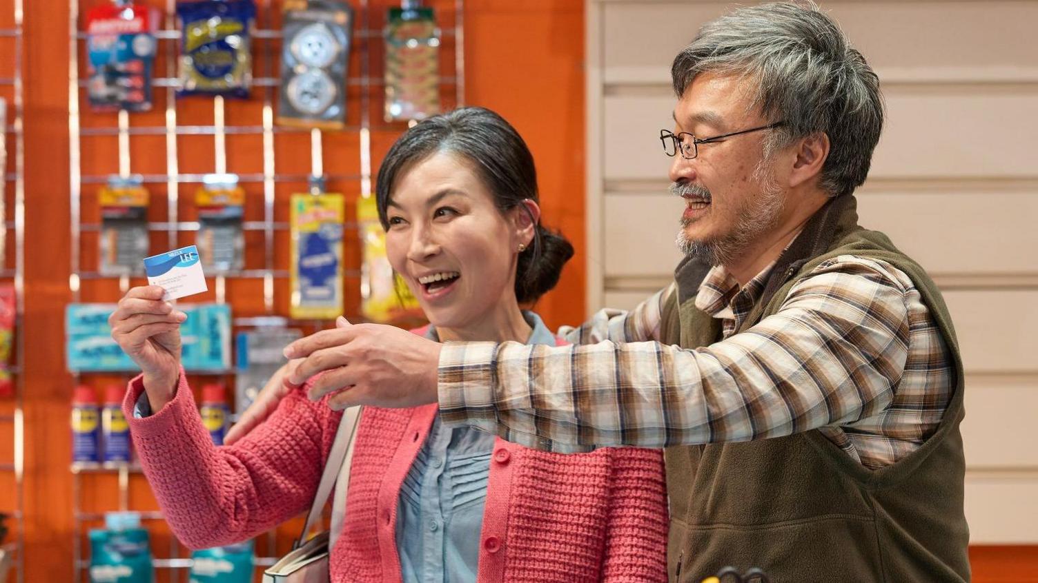 A man and a woman are behind a convenience store counter looking at a business card