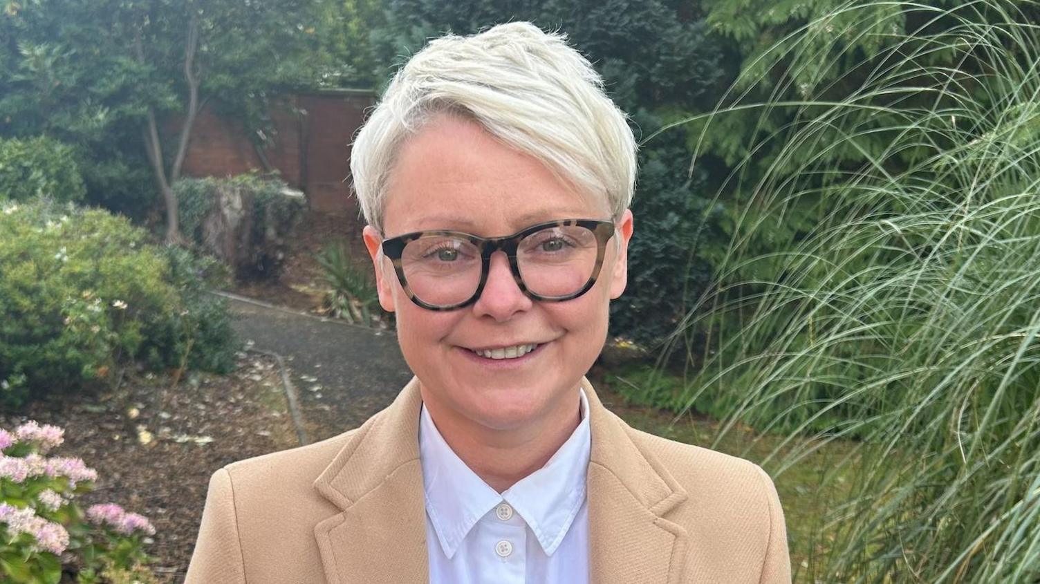 Karen McAlister is standing outside near a garden, she is wearing a cream coat and white shirt. Her hair is blonde and short, she is also wearing turtle pattern framed glasses.
