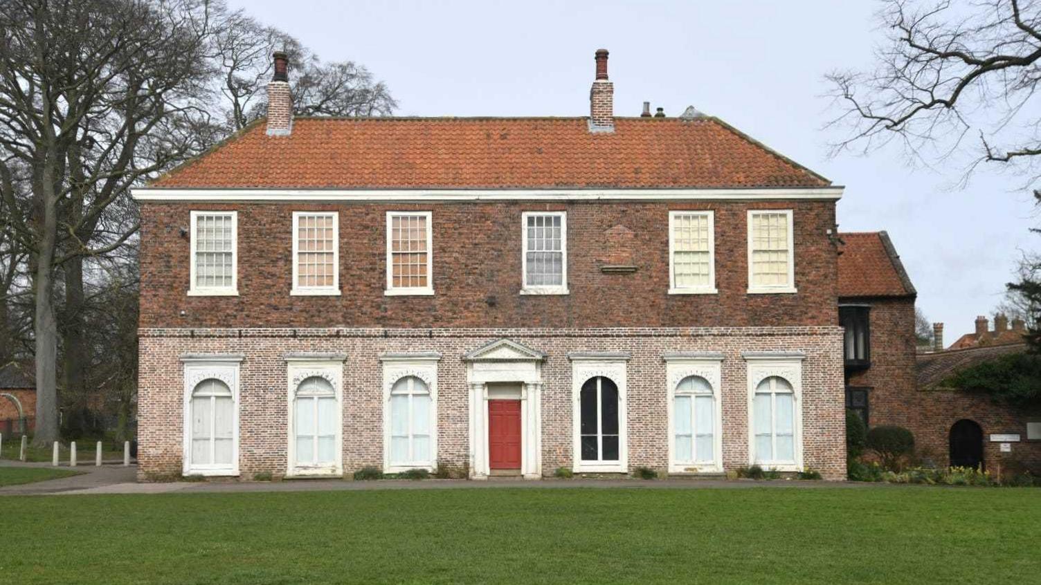 The grand Georgian frontage of the brick-built, 18th century Baysgarth House. The ground floor has a red door with white portico and three tall windows, with white frames, to either side.  The first floor has six tall windows and a small, bricked-up window. The orange-tiled roof has two chimney pots. The house stands in grounds with a green lawn and trees.