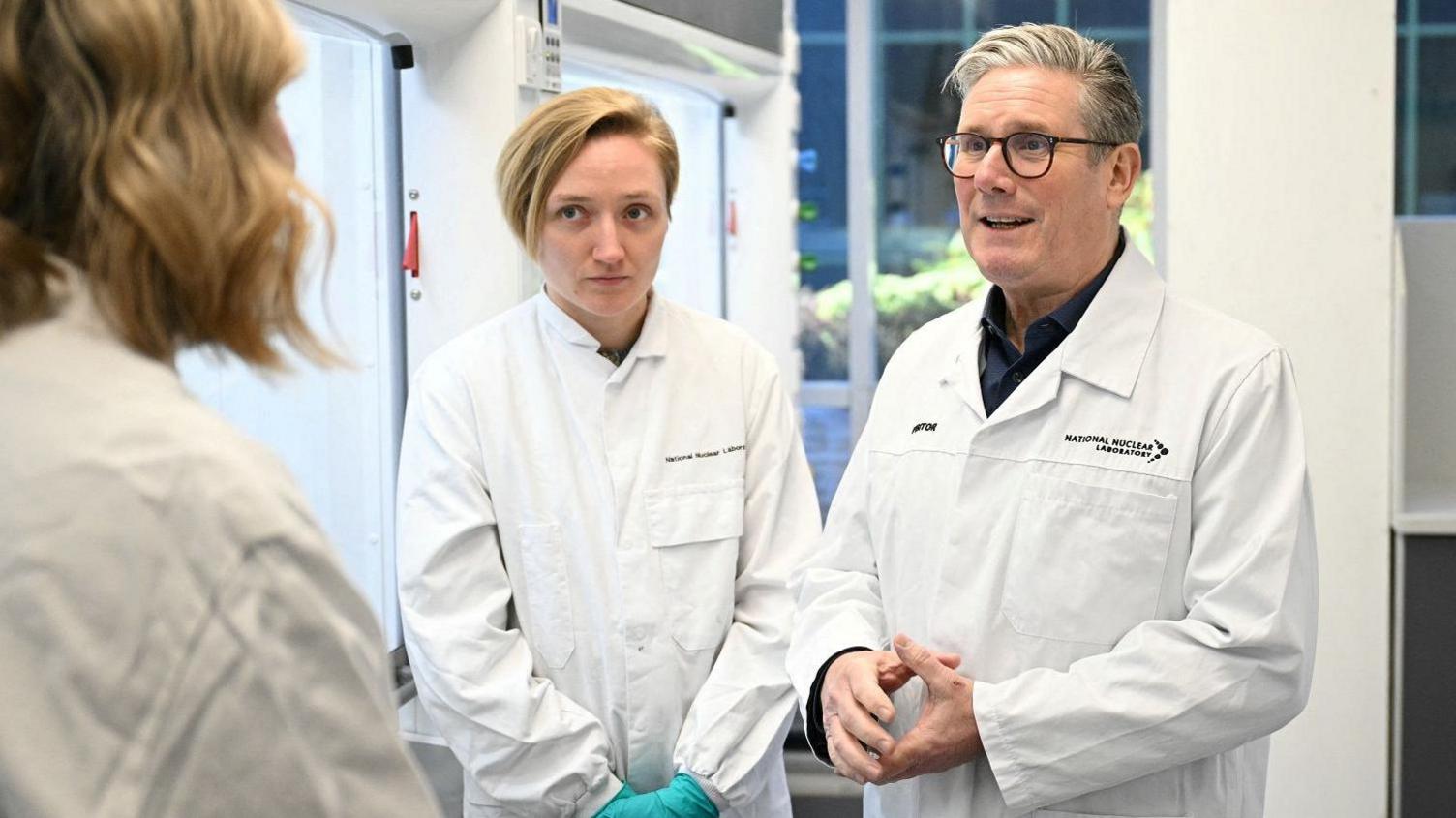 Prime Minister Sir Keir Starmer speaks with two female members of staff at the National Nuclear Laboratory faclity in Preston, Lancashire. They are all wearing white lab coats.