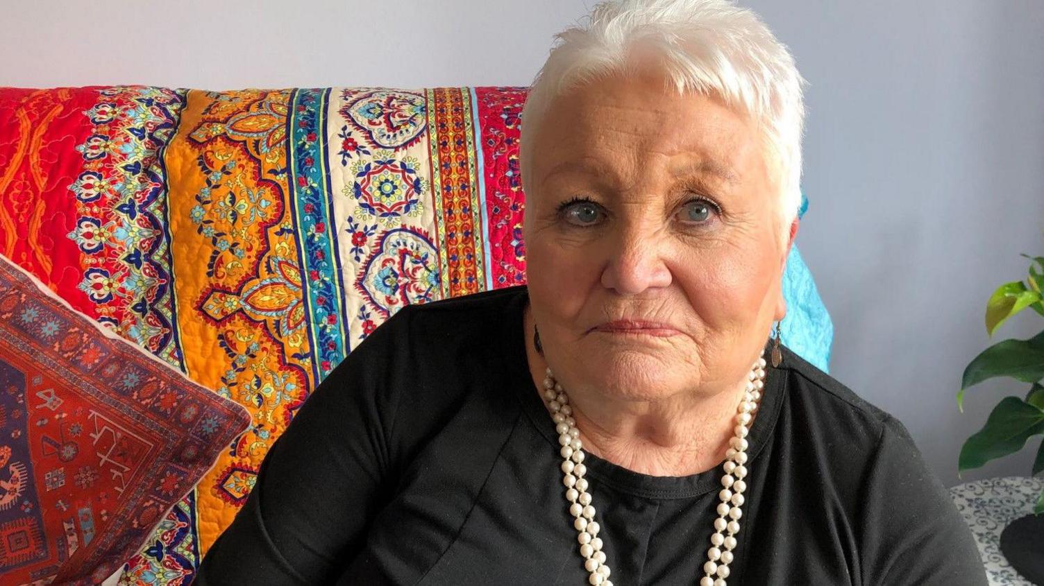 Pat Vinycomb looks at the camera. She has short, white hair and is wearing a black top with a string of white beads around her neck. Behind her on the chair is a colourful, patterned throw and cushions.