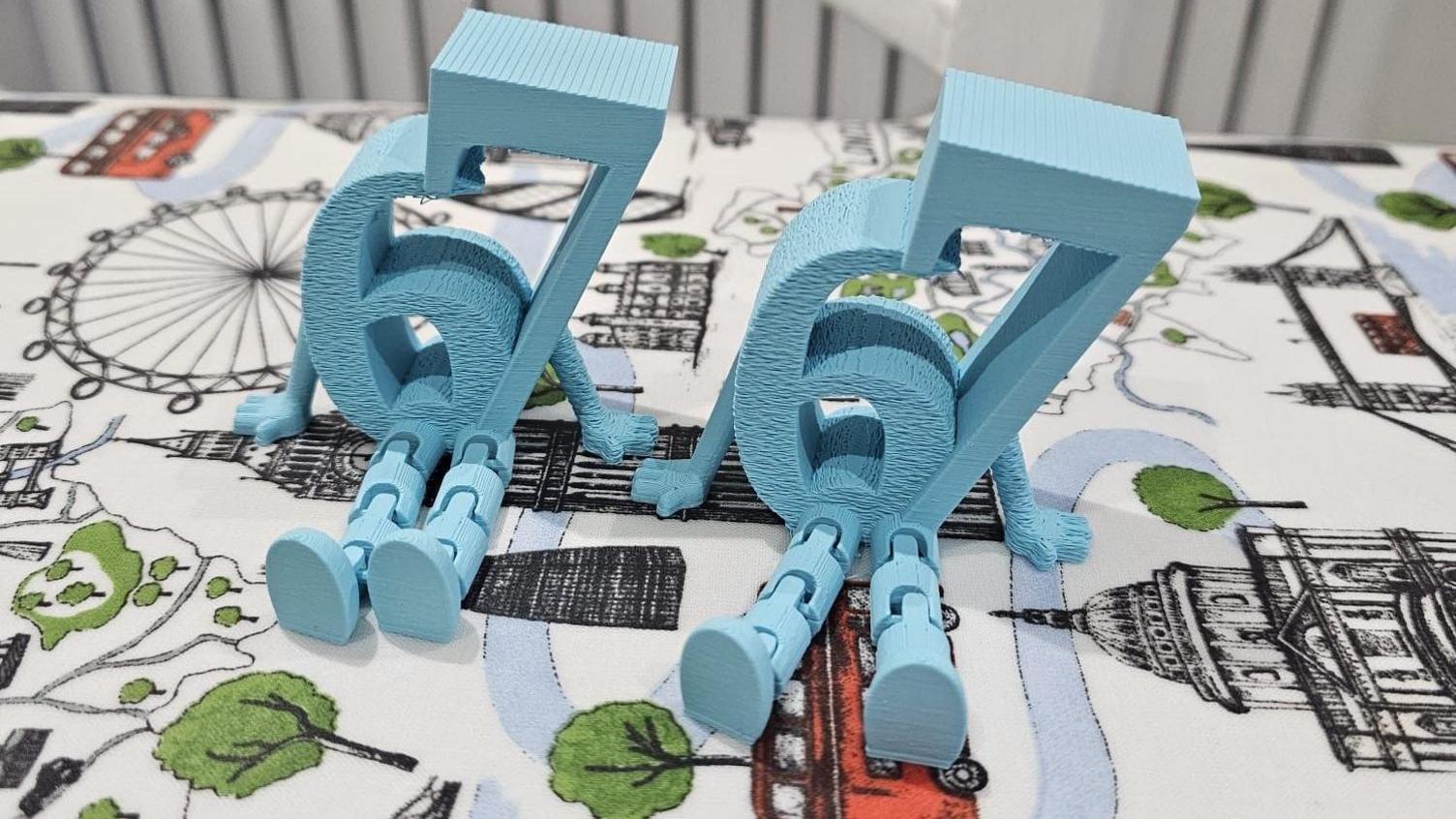 Two small plastic statues of the numbers '67', which have little arms and legs attached, are sat on a table covered in a printed white tablecloth.