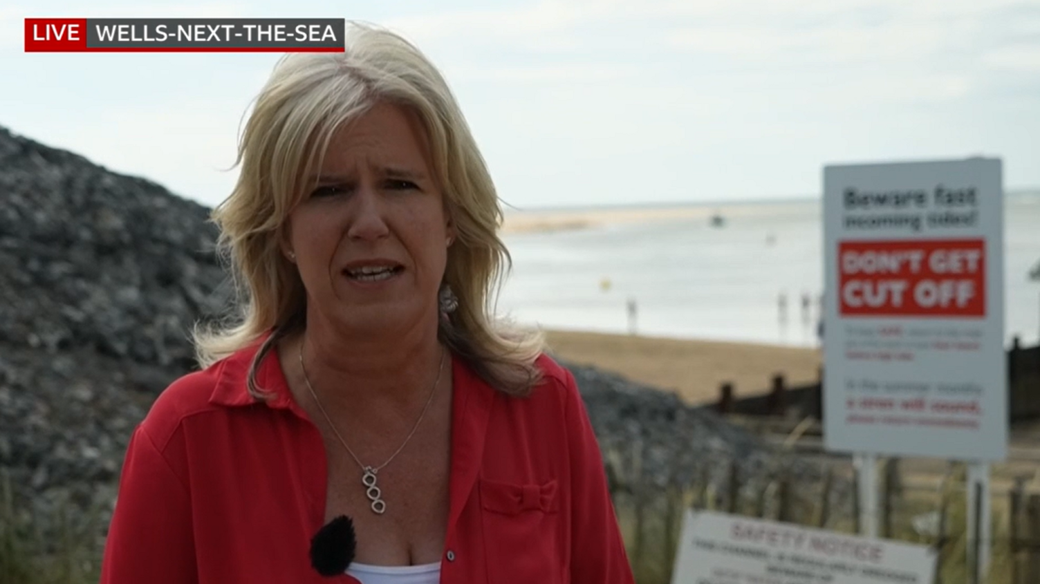 BBC reporter Debbie Tubby, who has shoulder-length blonde hair and is wearing a red jacket over a white top, reports live from a vantage point at the beach. A white and red sign is behind her, warning beach-goers of the dangers of the tide. Sand and water is even further in the background.