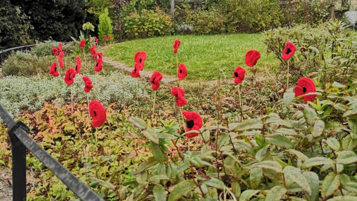 Knitted poppies on sticks in a garden beside the pavement.